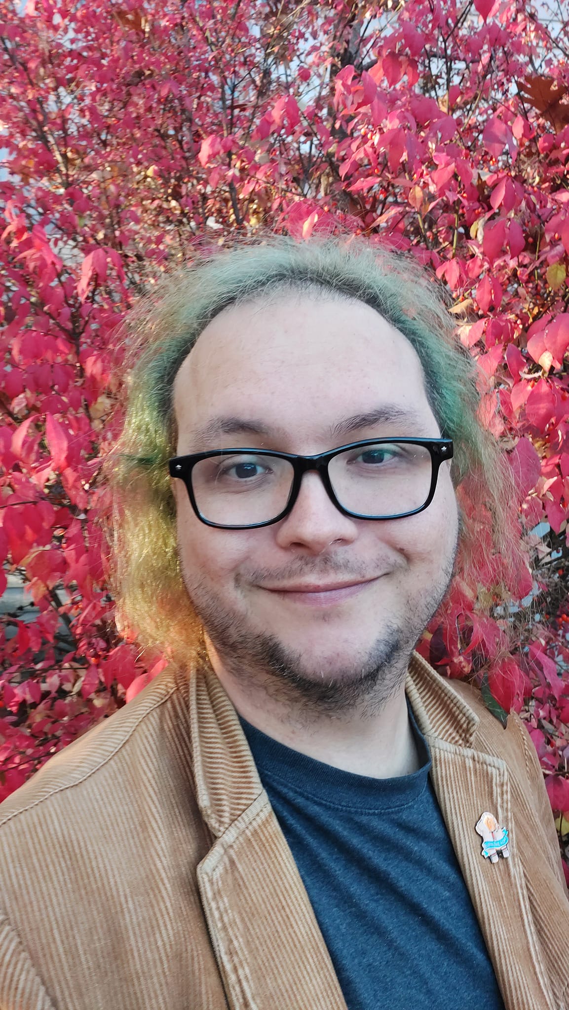 A man with green hair wearing a brown corderoy jacket stands in front of a bright red bush looking happy